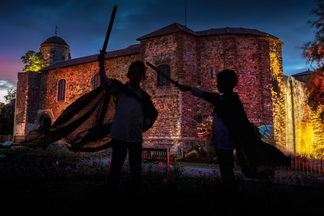 Two children in silhouette, dressed as wizards with capes and raised wands, appear to duel in front of a dramatically lit stone castle at dusk. The castle is illuminated in warm hues of yellow, orange, and purple, with a turret on the left and arched windows visible. A projection of a dragon’s head and wings is cast onto the castle wall, enhancing the magical atmosphere. The foreground is dark, with hints of garden flowers and a bench barely visible.