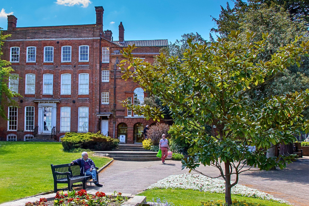 A historic brick building with multiple windows and chimneys stands at the end of a paved pathway, bordered by vibrant flower beds in full bloom. A large tree dominates the foreground, casting shade over part of the garden. One person sits on a bench near the flowers, while another walks toward the building. The scene is bathed in clear blue skies, suggesting a warm, pleasant day.